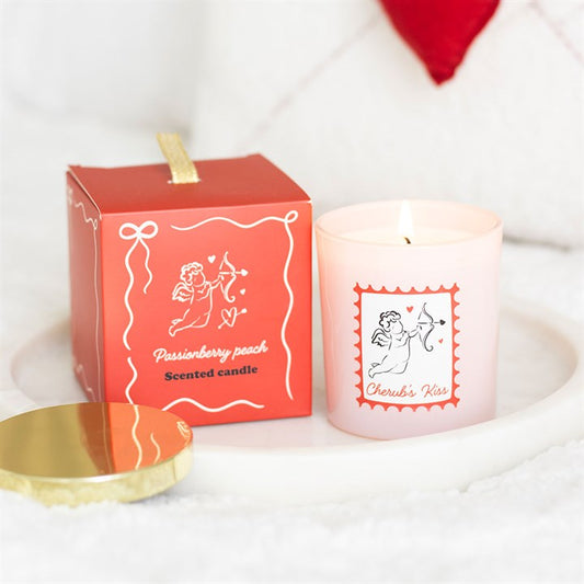 A lit Cherub’s Kiss scented candle featuring a cherub illustration rests on a tray next to its matching red box, labelled “Passionberry Peach Scented Candle.” Its romantic fragrance is ideal for Valentine’s Day.