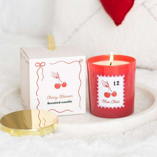 A red cherry blossom candle with a burning flame rests next to its white box and gold lid. Both are adorned with cherry illustrations and French text, making an ideal self-care or romantic gift on a cosy white surface.