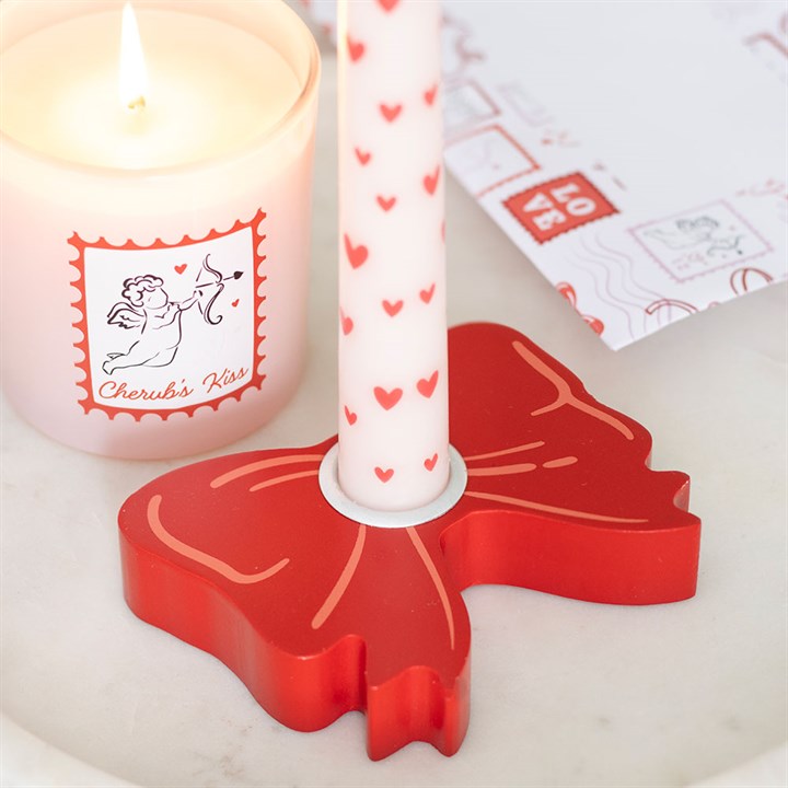 A lit candle in a glass adorned with a cherub label, a white taper candle decorated with red hearts in a red bow holder, and stationery featuring red love-themed designs on white marble—ideal for Valentine’s Day.