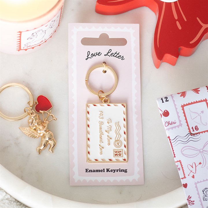A gold enamel love letter keyring sits on a pink card labelled Love Letter Enamel Keyring. Nearby are a cupid keyring, candle, red tray, and an envelope decorated with heart motifs—a charming Valentine’s Day keepsake.