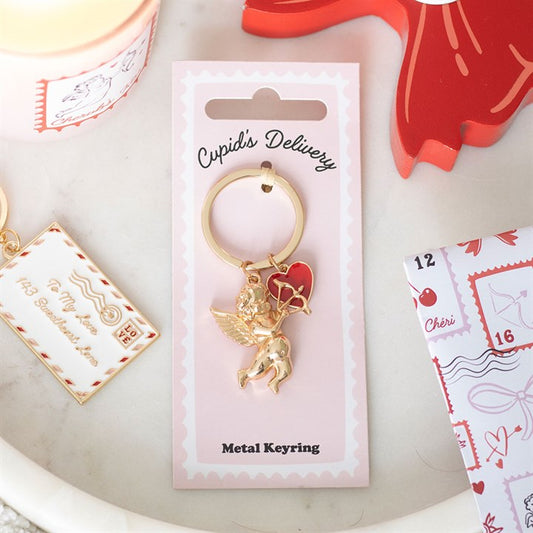 A gold cupid keyring featuring a winged cupid, red heart charm, and gold heart outline, displayed on pink Cupids Delivery packaging. Surrounded by decorative post-themed items—a lovely Valentine’s Day gift.