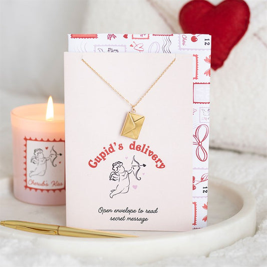 A gold love letter locket necklace sits on a card reading “Cupid’s delivery. Open envelope to read secret message.” Nearby are a lit, romantic candle, pen, and red heart cushion—ideal as a Valentine’s Day gift.
