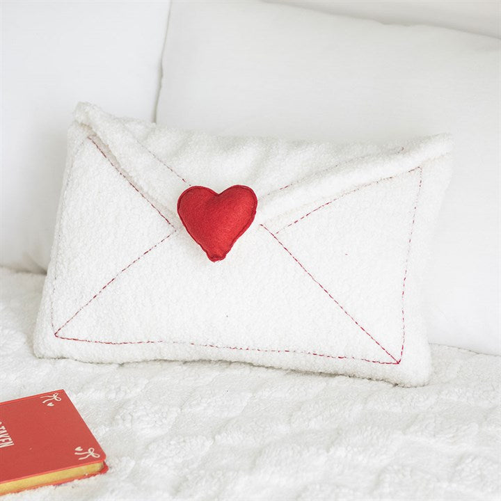 A plush cushion shaped like a white envelope with red stitching, a red heart seal, and an embroidered message rests on a white bouclé cushion bedspread beside a red book. White pillows are in the background.