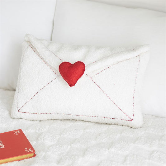 A plush cushion shaped like a white envelope with red stitching, a red heart seal, and an embroidered message rests on a white bouclé cushion bedspread beside a red book. White pillows are in the background.