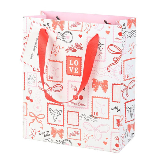 A white Valentine’s Day gift bag decorated with red and pink LOVE-themed designs—bows, hearts, and stamps. It has red ribbon handles, a matching gift tag, and a pale pink interior.