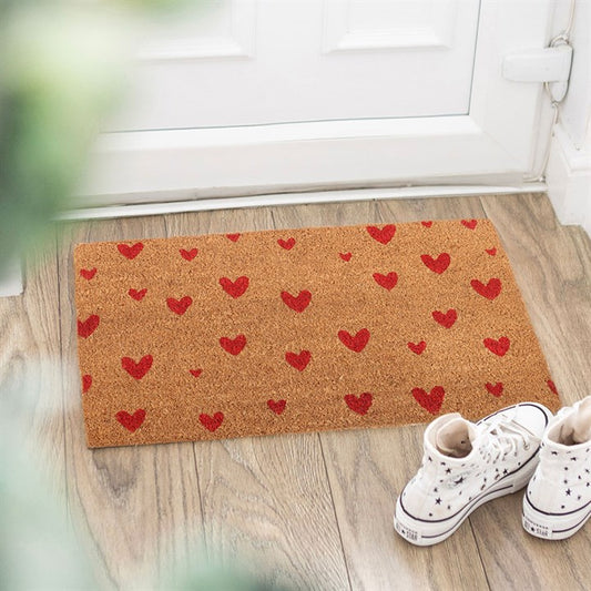 A brown doormat shaped like a love heart, scattered with red hearts, sits on a light wooden floor by a white door. White high-top trainers with blue stars are placed to the right, while blurred green leaves frame the romantic doorstep décor.