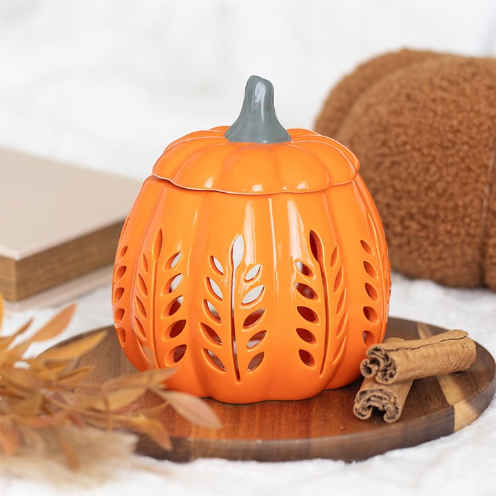 A ceramic orange pumpkin oil burner with leaf-shaped cut-outs sits on a wooden tray. Cinnamon sticks, dried leaves, a book, and a brown fluffy cushion complete this cosy autumn scene on a white textured surface.