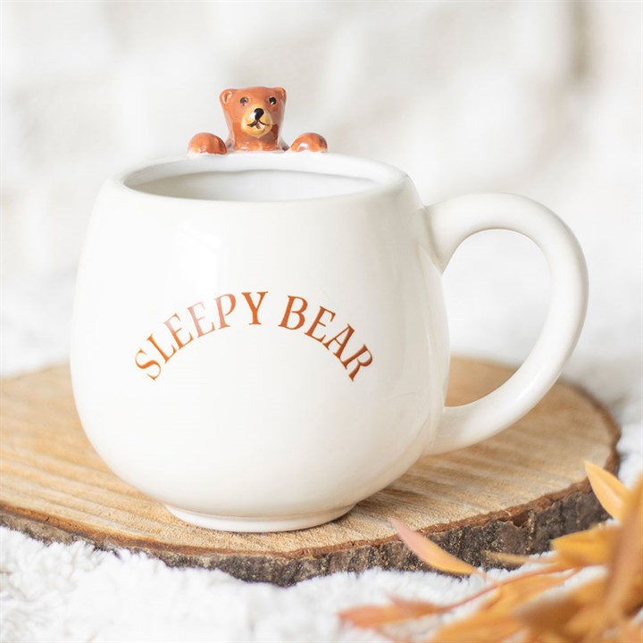 A Sleepy Bear mug with orange lettering rests on a wooden coaster. A tiny bear figurine peeks inside the ceramic mug, which is suitable for handwashing only. Soft white fabric and dried leaves surround it for a cosy touch.