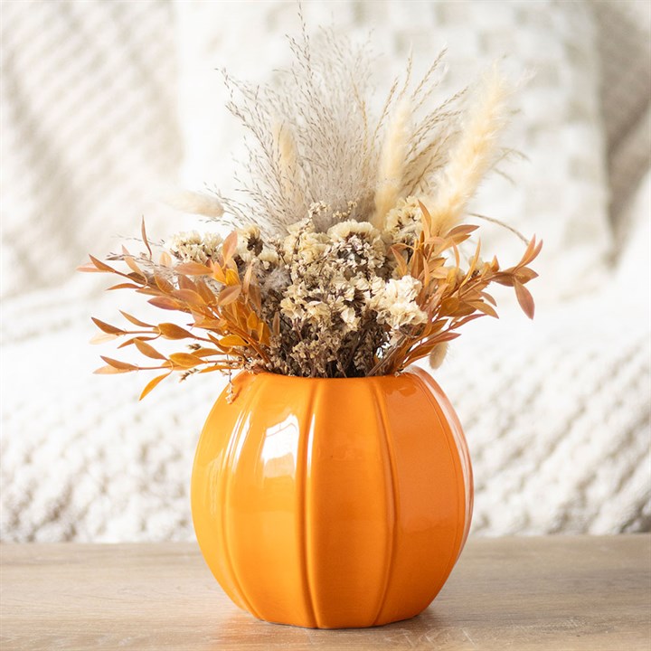 An orange, pumpkin-shaped ceramic vase holds dried grasses and flowers, including beige pampas grass and orange leaves, set on a wooden surface with a textured white blanket in the background—perfect for autumn décor.