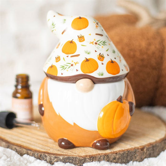 A ceramic gnome figurine with a white beard, holding a pumpkin. Its tall hat features a charming pumpkin print and autumn leaves. It sits on a wooden slice beside an amber fragrance oil bottle, evoking a cosy autumn theme.