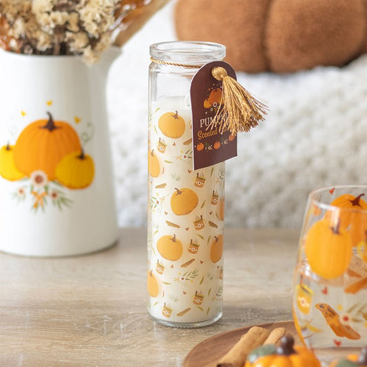 A tall prayer candle featuring an autumnal design of orange pumpkins and cinnamon sticks stands on a wooden table. A brown tag with a gold tassel hangs nearby, next to a vase with a pumpkin print and a glass decorated with orange designs.