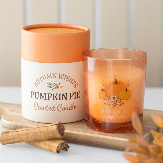 An orange pumpkin pie candle in a glass jar sits beside its matching autumn-themed container, perfect as an autumn gift for cosy evenings. Cinnamon sticks and blurred autumn leaves complete the warm scene.