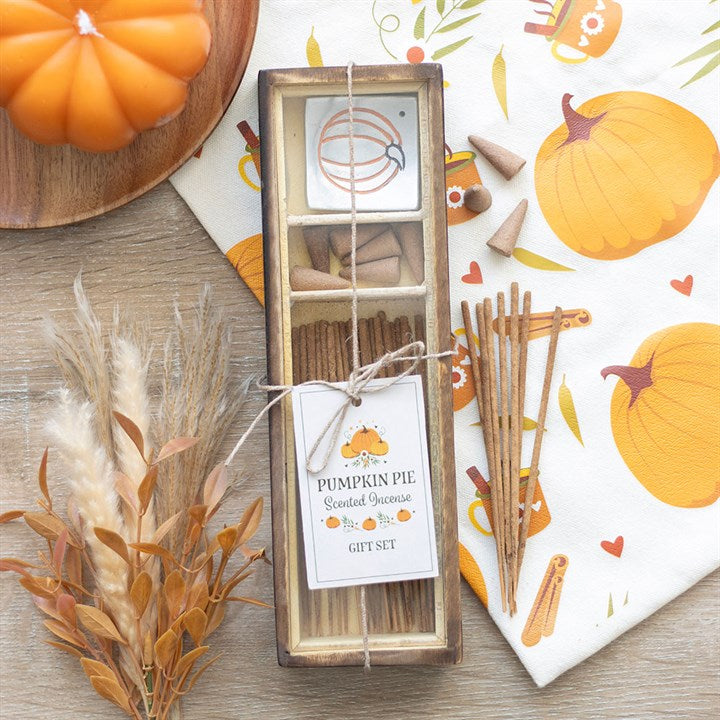 A pumpkin pie-scented incense gift set rests on a wooden surface, featuring sticks, cones, and a ceramic holder. Autumn-themed fragrances fill the air amidst cosy décor, dried plants, and a napkin illustrated with a pumpkin.