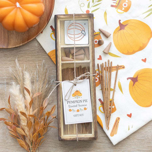 A pumpkin pie-scented incense gift set rests on a wooden surface, featuring sticks, cones, and a ceramic holder. Autumn-themed fragrances fill the air amidst cosy décor, dried plants, and a napkin illustrated with a pumpkin.