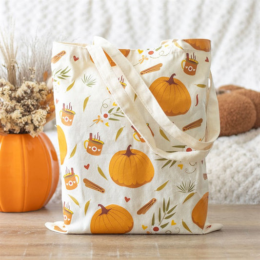 A beige polycotton tote bag with an autumn pumpkin design featuring cinnamon sticks, small hearts, and steaming mugs rests on a wooden table. Beside it is an orange vase filled with dried flowers against a blurred, cosy backdrop.