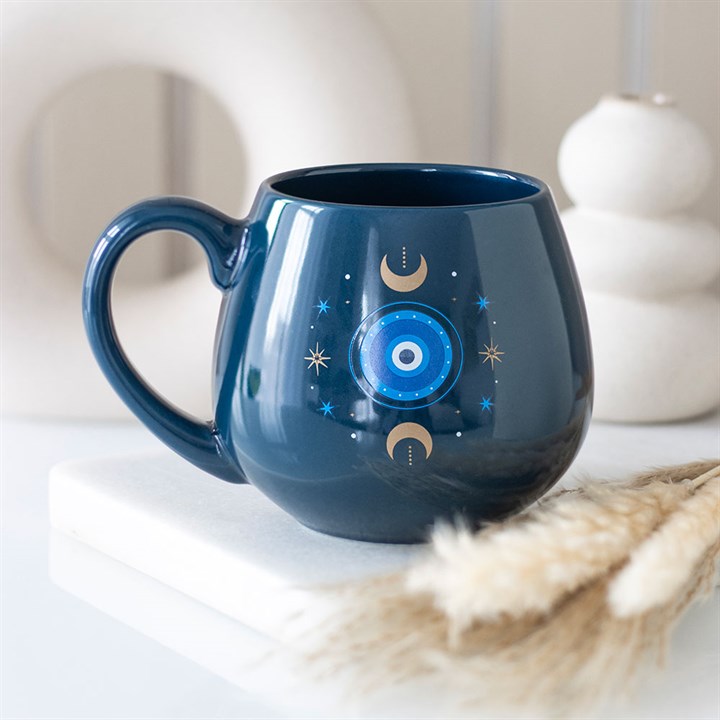A round, dark blue celestial mug with a large handle sits on a white surface. The mug features moons, stars, and a blue concentric circle pattern. Beige decorative objects and dried grasses blur in the background. Rewritten in British English: A round, dark blue celestial mug with a large handle rests on a white surface. The mug is decorated with moons, stars, and a pattern of blue concentric circles. In the background, beige decorative items and dried grasses appear blurred.