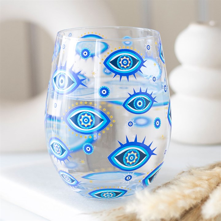 A clear glass tumbler decorated with blue and gold all-seeing eye designs sits on a white surface, bringing an otherworldly style to décor softly blurred in the background.