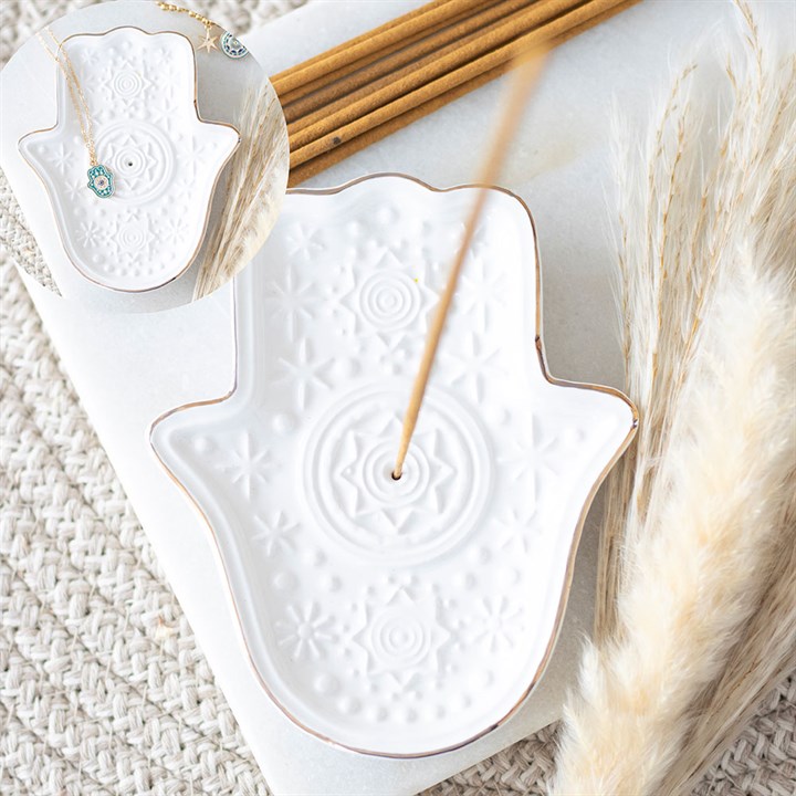 A white, patterned ceramic Hamsa incense holder with a burning stick rests on a textured surface beside dried pampas grass. An inset shows a turquoise eye necklace displayed on the trinket dish.