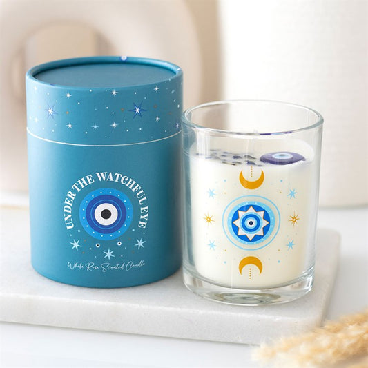 A white, rose-scented all-seeing eye candle in a glass jar adorned with celestial designs sits beside its blue, star-patterned box, making a perfect celestial gift beneath the text UNDER THE WATCHFUL EYE.