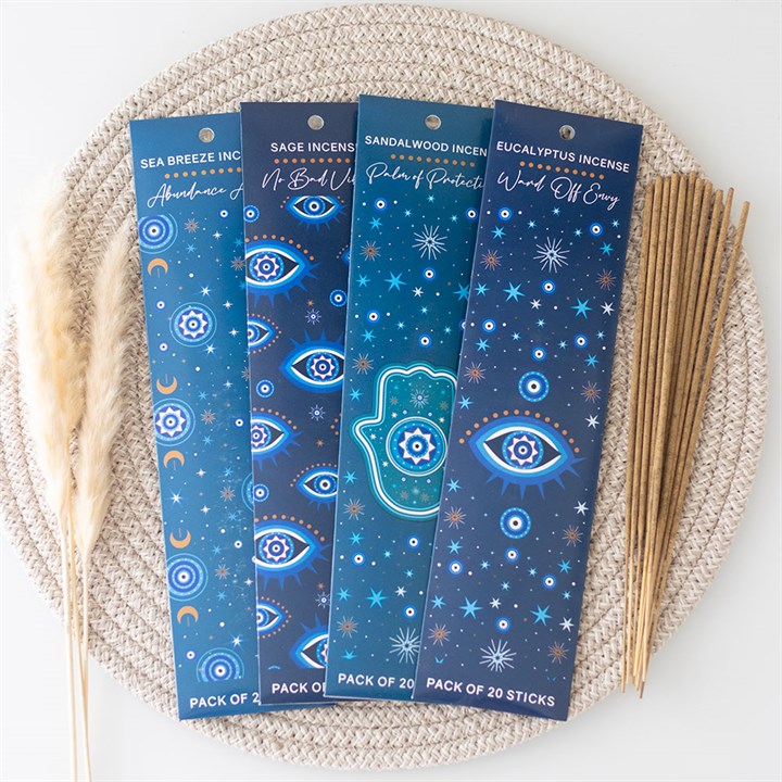 Four blue incense stick packs adorned with celestial designs of mystical eyes and stars rest on a woven placemat. Each offers calming fragrances. Loose incense sticks and dried grass are arranged nearby.
