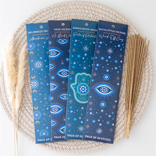 Four blue incense stick packs adorned with celestial designs of mystical eyes and stars rest on a woven placemat. Each offers calming fragrances. Loose incense sticks and dried grass are arranged nearby.