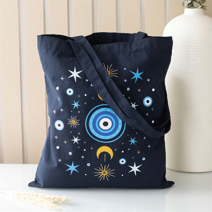 A navy blue tote bag with a long strap features a celestial pattern and a subtle all-seeing eye design, evoking a spiritual style. The bag rests on a white surface next to a textured white vase.