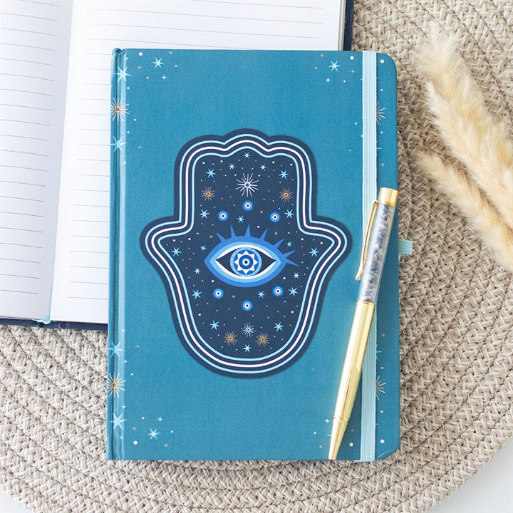 A turquoise notebook featuring a Hamsa hand design with an eye at the centre lies on a woven mat. A pen filled with crystal chips rests on top, while pampas grass and an open lined notebook are partially visible nearby.