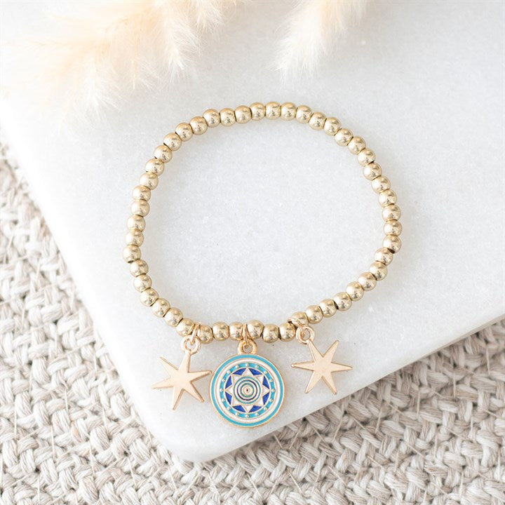A gold beaded charm bracelet featuring two gold star charms and a blue-and-gold circular mandala, displayed on a white stone slab atop woven beige fabric, with soft cream dried flowers in the corner.
