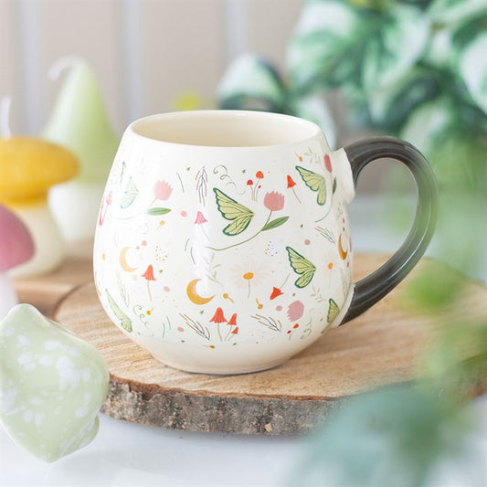 A round ceramic mug with a black handle rests on a wooden slice coaster. The whimsical green moths, orange flowers, and yellow moons lend a garden fairy charm, surrounded by blurred plants and mushroom décor.