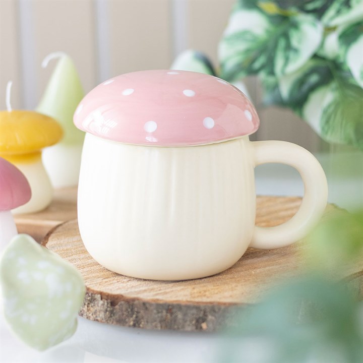 A cream-coloured ceramic mug with a pink, white-spotted mushroom cap lid sits on a wooden coaster, surrounded by woodland magic—decorative mushroom candles and green leafy plants in the background.