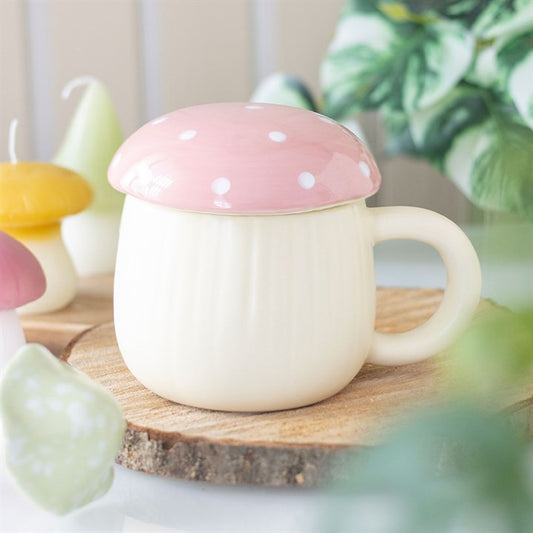 A cream-coloured ceramic mug with a pink, white-spotted mushroom cap lid sits on a wooden coaster, surrounded by woodland magic—decorative mushroom candles and green leafy plants in the background.