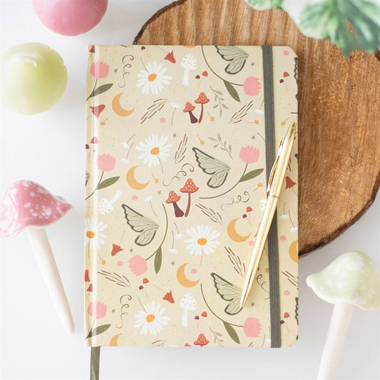 A beige notebook featuring a garden fairy print and whimsical illustrations rests on a wooden surface. A gold pen lies atop it, while pastel candles and ceramic mushrooms surround this perfect companion for creative journalling.