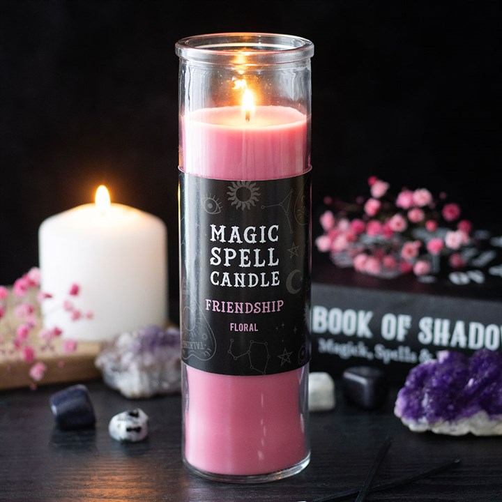 A lit Friendship Magic Spell candle stands on a dark table with crystals, a white prayer candle, pink flowers, and a “Book of Shadows” in the background, creating a mystical ambience.