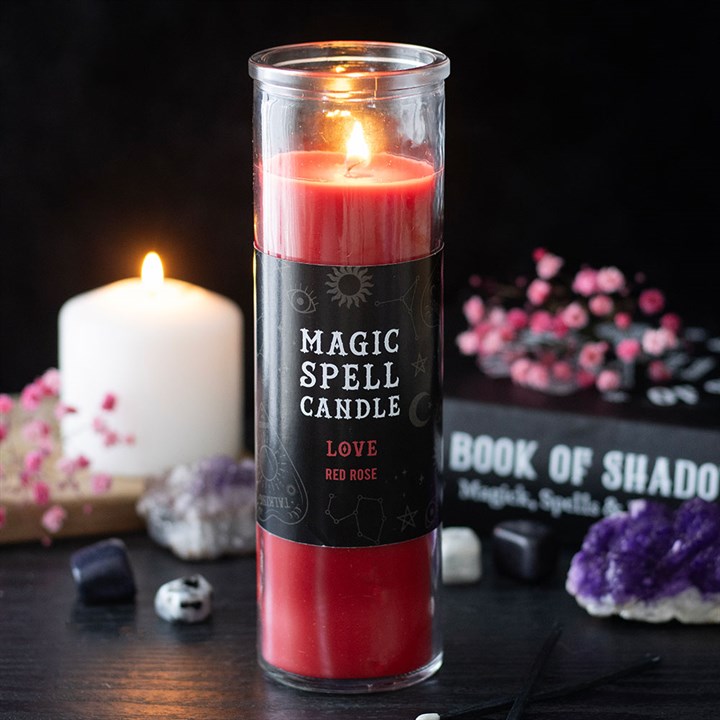 A lit Love Magic Spell candle labelled “Love: Red Rose” stands on a dark table, ideal for romantic rituals. Nearby are crystals, pink flowers, and a Book of Shadows amidst mystical décor, evoking a rose scent.