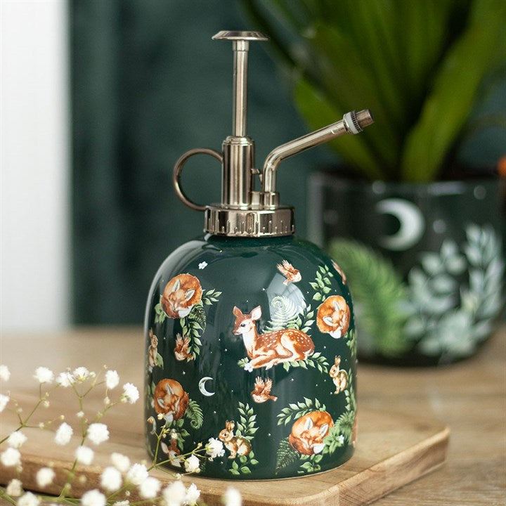 A ceramic plant mister with a metal pump and spout, featuring a woodland design of painted foxes and flowers, sits on a wooden surface. Gardening accessories and a leafy plant in a matching pot are blurred in the background.