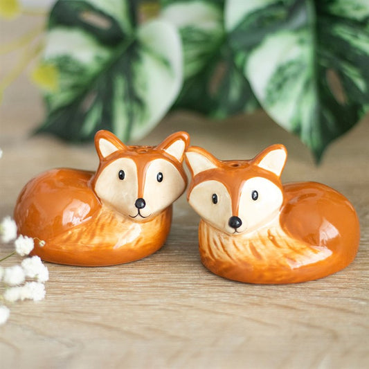 Two ceramic fox salt and pepper shakers sit side by side on a wooden surface, adding quirky charm to the kitchen. Green leafy plants are blurred in the background behind their orange and white fur with black eyes and noses.
