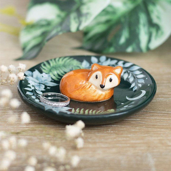 A charming ceramic trinket dish with painted green leaves holds a cute orange fox figurine and a sparkling silver ring. Blurred flowers and green foliage adorn the background on a wooden surface.