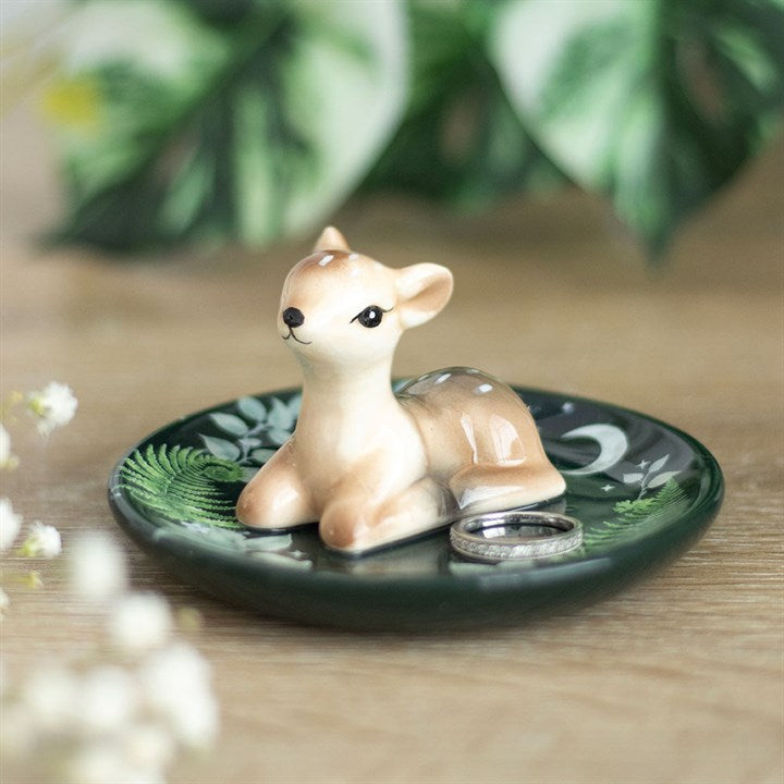 A small ceramic fawn figurine sits on a dark green ceramic trinket dish with leafy patterns, holding two silver rings beside it—perfect for animal lovers and nature enthusiasts. Blurred greenery and white flowers complete the scene. British English revision: A small ceramic fawn ornament rests on a dark green ceramic trinket dish adorned with leafy patterns, holding two silver rings beside it—ideal for animal lovers and nature enthusiasts. Soft-focus greenery and white flowers complete the scene.