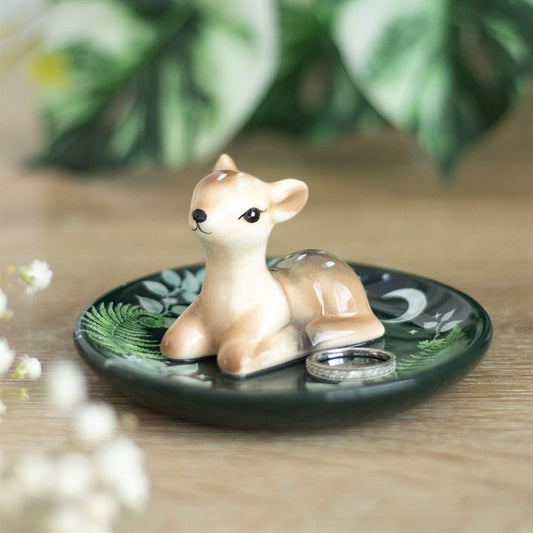 A small ceramic fawn figurine sits on a dark green ceramic trinket dish with leafy patterns, holding two silver rings beside it—perfect for animal lovers and nature enthusiasts. Blurred greenery and white flowers complete the scene. British English revision: A small ceramic fawn ornament rests on a dark green ceramic trinket dish adorned with leafy patterns, holding two silver rings beside it—ideal for animal lovers and nature enthusiasts. Soft-focus greenery and white flowers complete the scene.
