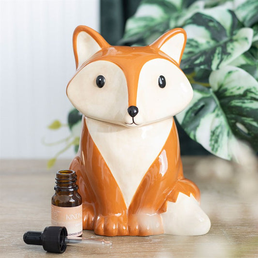 A ceramic, fox-shaped oil burner sits on a wooden surface beside an open essential oil bottle and dropper. Green, leafy houseplants blur in the background, highlighting the fox’s charming, wide-eyed woodland creature appeal.