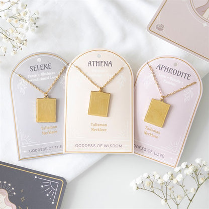 Three gold-toned Talisman necklaces labelled SELENE, ATHENA, and APHRODITE showcase attributes such as peace and beauty, capturing the essence of goddess energy. Surrounded by white flowers on a pastel background, a glimpse of another card peeps from the top right.