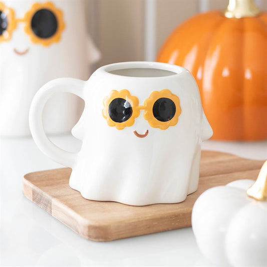 A ghost-shaped ceramic mug with orange flower-shaped sunglasses and a smiling face sits on a wooden board among pumpkin decorations—perfect quirky homeware or a fun Halloween gift.