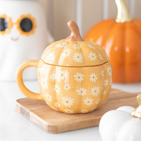 A quirky piece of homeware, this orange pumpkin-shaped mug with a lid features white daisy patterns and sits on a wooden board amid pumpkin and ghost decorations—perfect for your autumn drinkware collection.