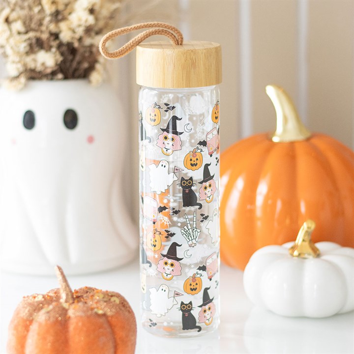 A clear Halloween water bottle with a bamboo lid and rope handle features a spooky design of witches, black cats, ghosts, pumpkins, and bones. In the background are a ghost vase and two pumpkins—one orange, one white.