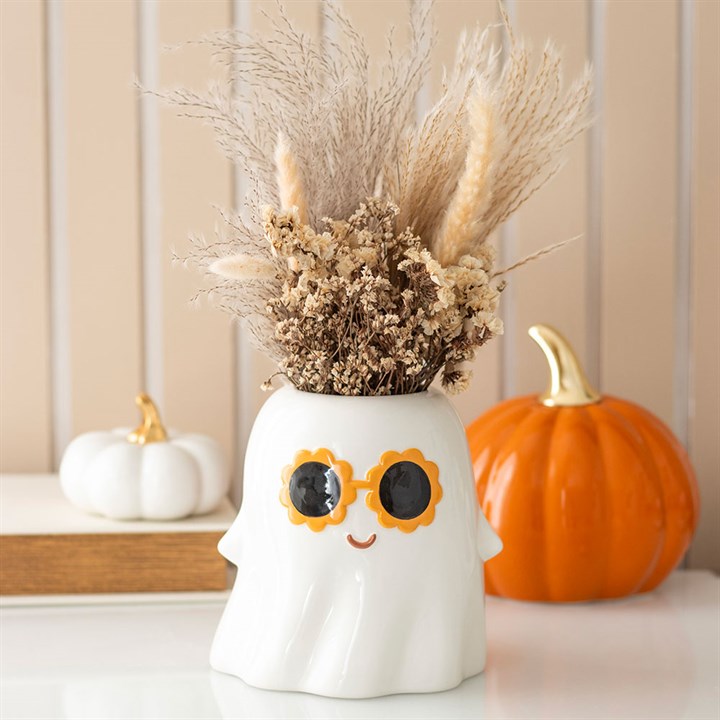 A ceramic ghost vase sporting orange-rimmed sunglasses and a smile holds dried grasses—perfect for spooky home décor. Behind it are two pumpkins, one white with a gold stem and one orange, set against a beige striped background.
