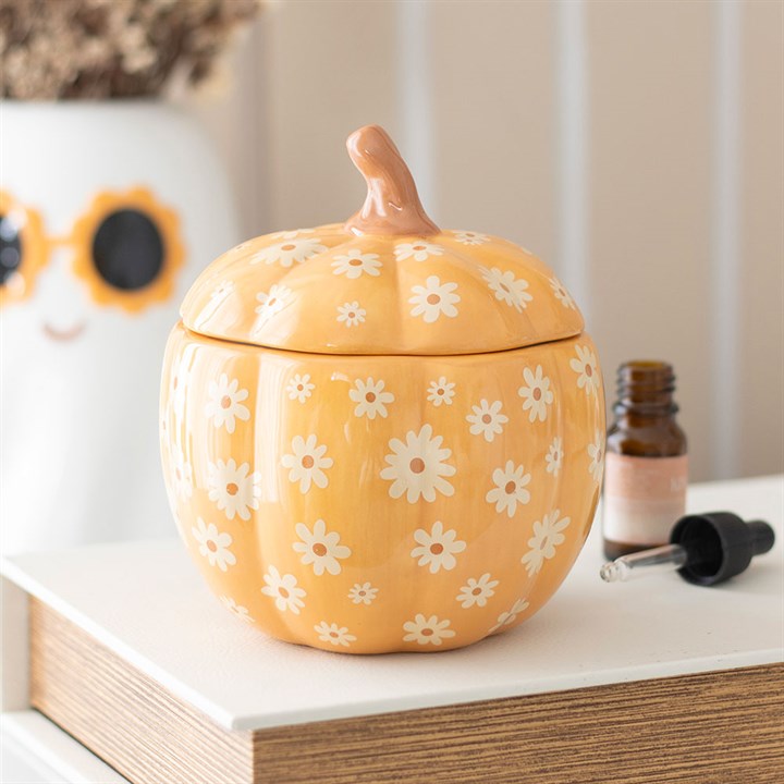 A ceramic, pumpkin-shaped oil burner with white daisy patterns sits on a table. Its lid has a brown stem—perfect Hallowe’en décor—alongside a dropper bottle and a white vase with a yellow flower and smiling face.