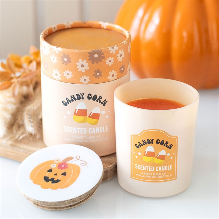 A cream-coloured, candy corn-scented candle in a glass jar sits beside its matching, floral-decorated box. The lid, featuring a smiling pumpkin, rests nearby—perfect for adding an autumnal aroma to Hallowe’en parties.