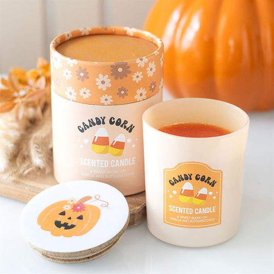 A cream-coloured, candy corn-scented candle in a glass jar sits beside its matching, floral-decorated box. The lid, featuring a smiling pumpkin, rests nearby—perfect for adding an autumnal aroma to Hallowe’en parties.