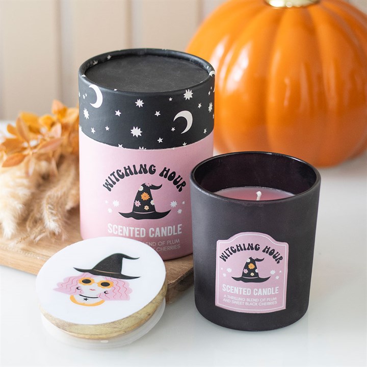 A pink-and-black Witching Hour candle with an enchanting aroma of plum and black cherry sits beside its starry box and witchs hat label; a pumpkin and dried leaves add festive charm in the background.