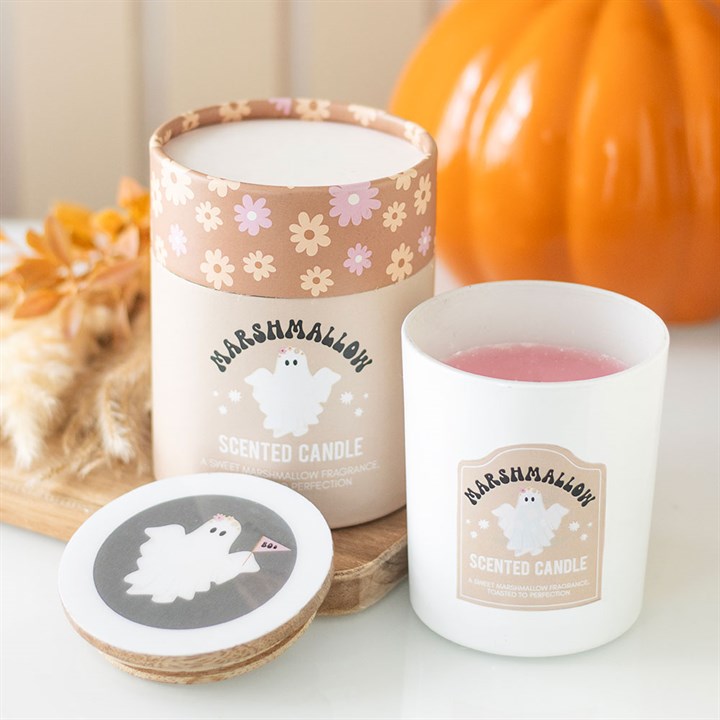 A white ghost candle in a glass jar labelled “Marshmallow Scented Candle” features a cartoon ghost; perfect for cosy autumn evenings, with its box and lid matching the design. A pumpkin and dried flowers are softly blurred in the background.